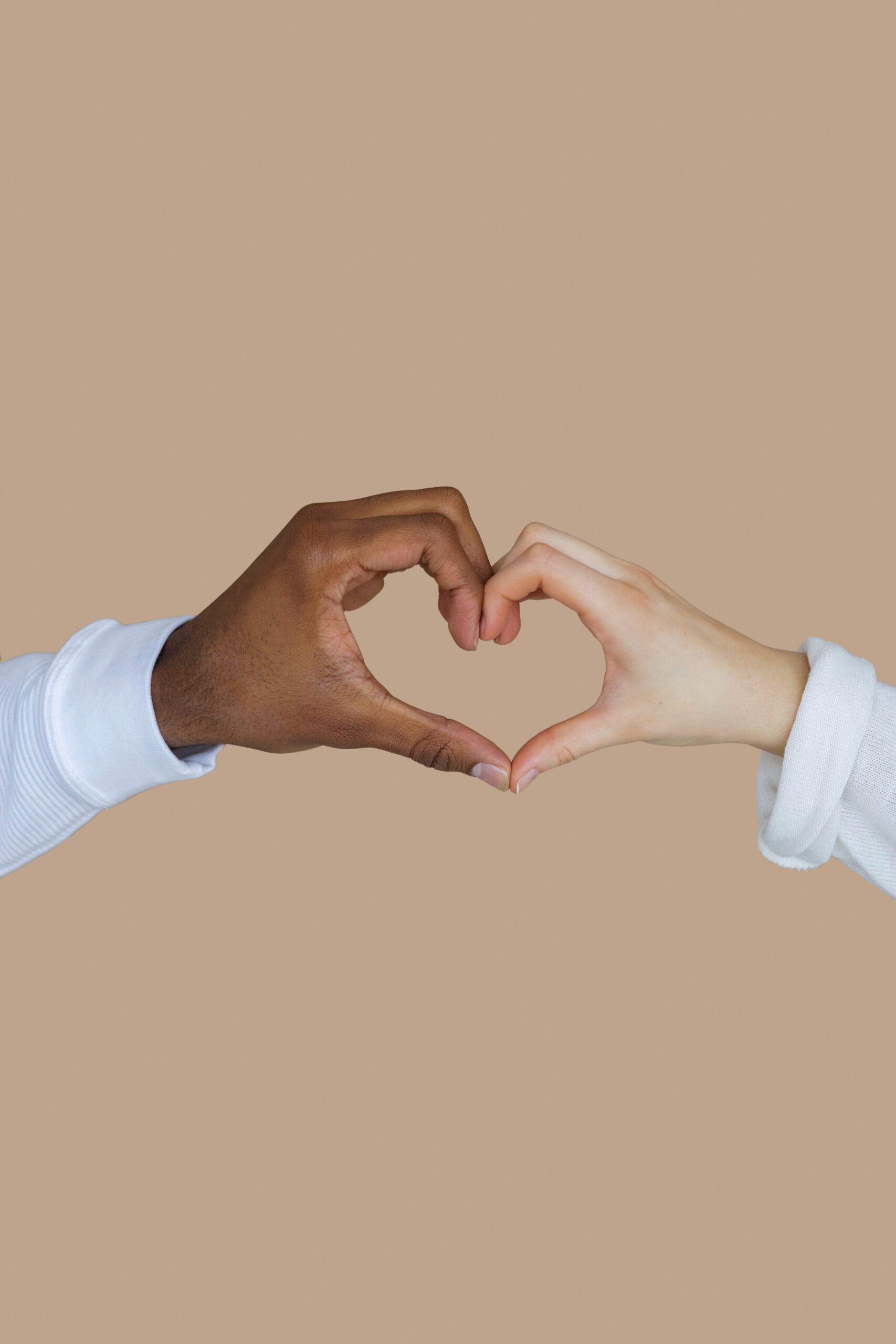 Two people with different skin tones form a heart shape with their hands against a neutral beige background.