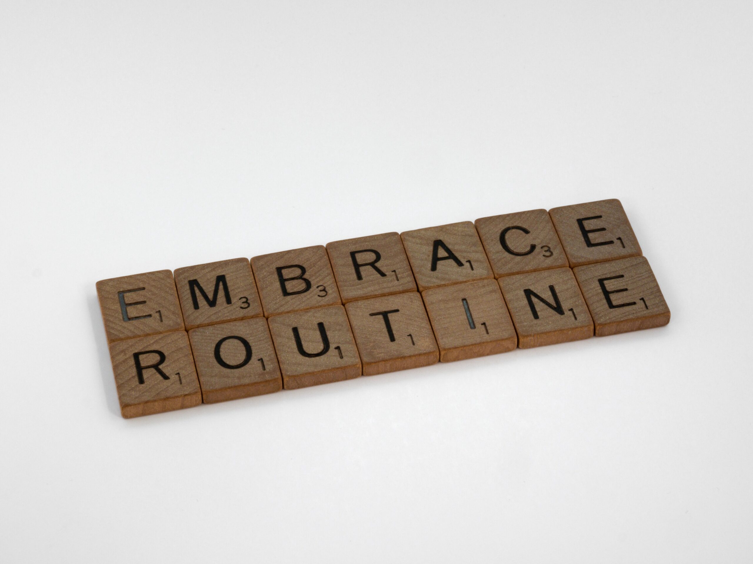 Wooden letter tiles arranged on a white background spell “EMBRACE ROUTINE.”