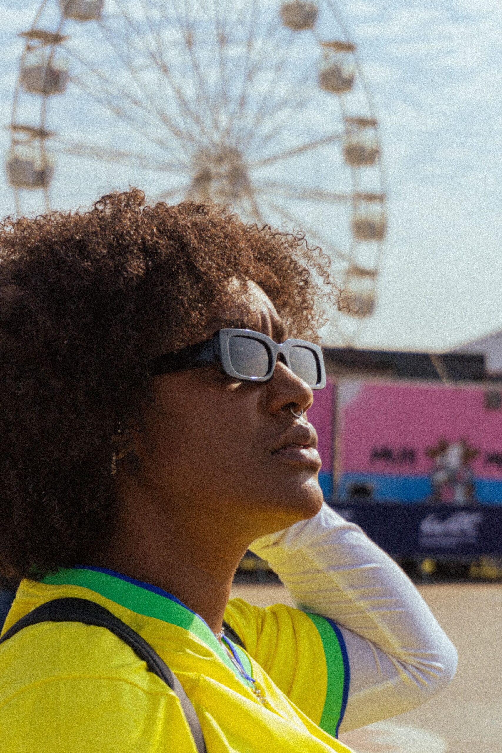 Person with curly hair and sunglasses wearing a yellow shirt, with a Ferris wheel in the background.