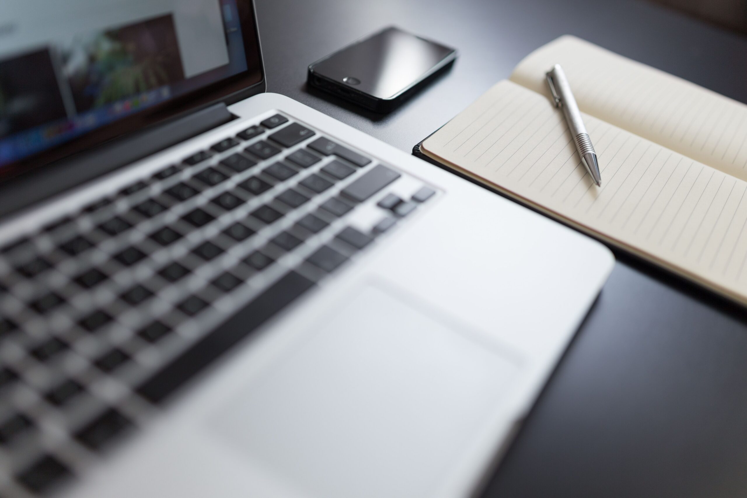 Laptop, smartphone, and open notebook with a pen arranged on a workplace desk.