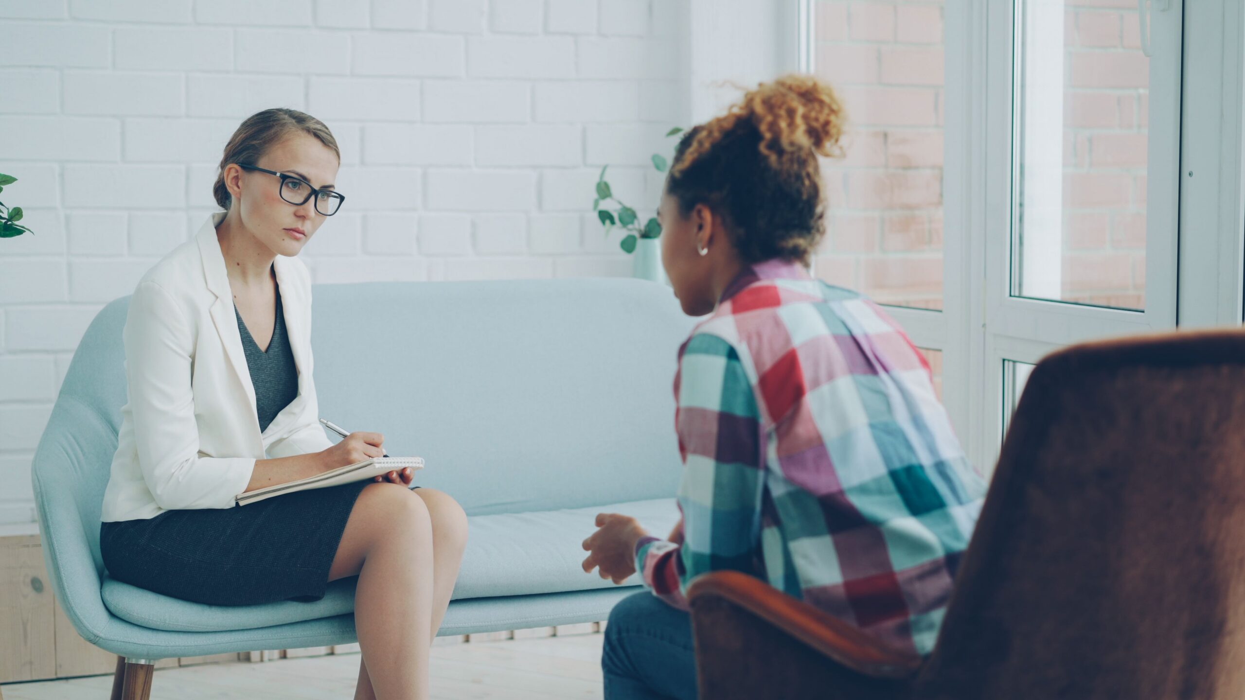 Two women sit facing each other in a bright office, one listening with a pen and notebook as the other speaks.